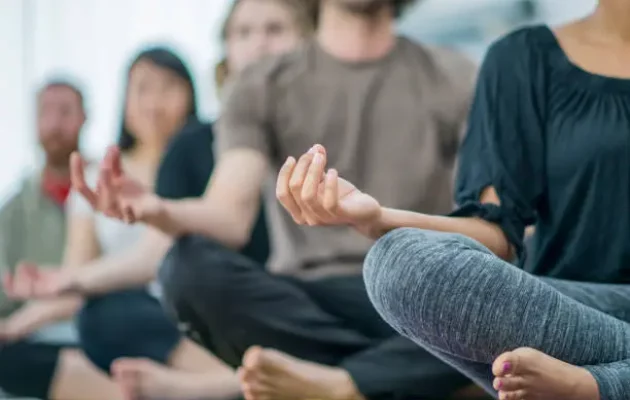 A multi-ethnic group of young adults are indoors in a fitness center. They are wearing casual clothing. They are meditating while sitting on the floor in this angled shot.